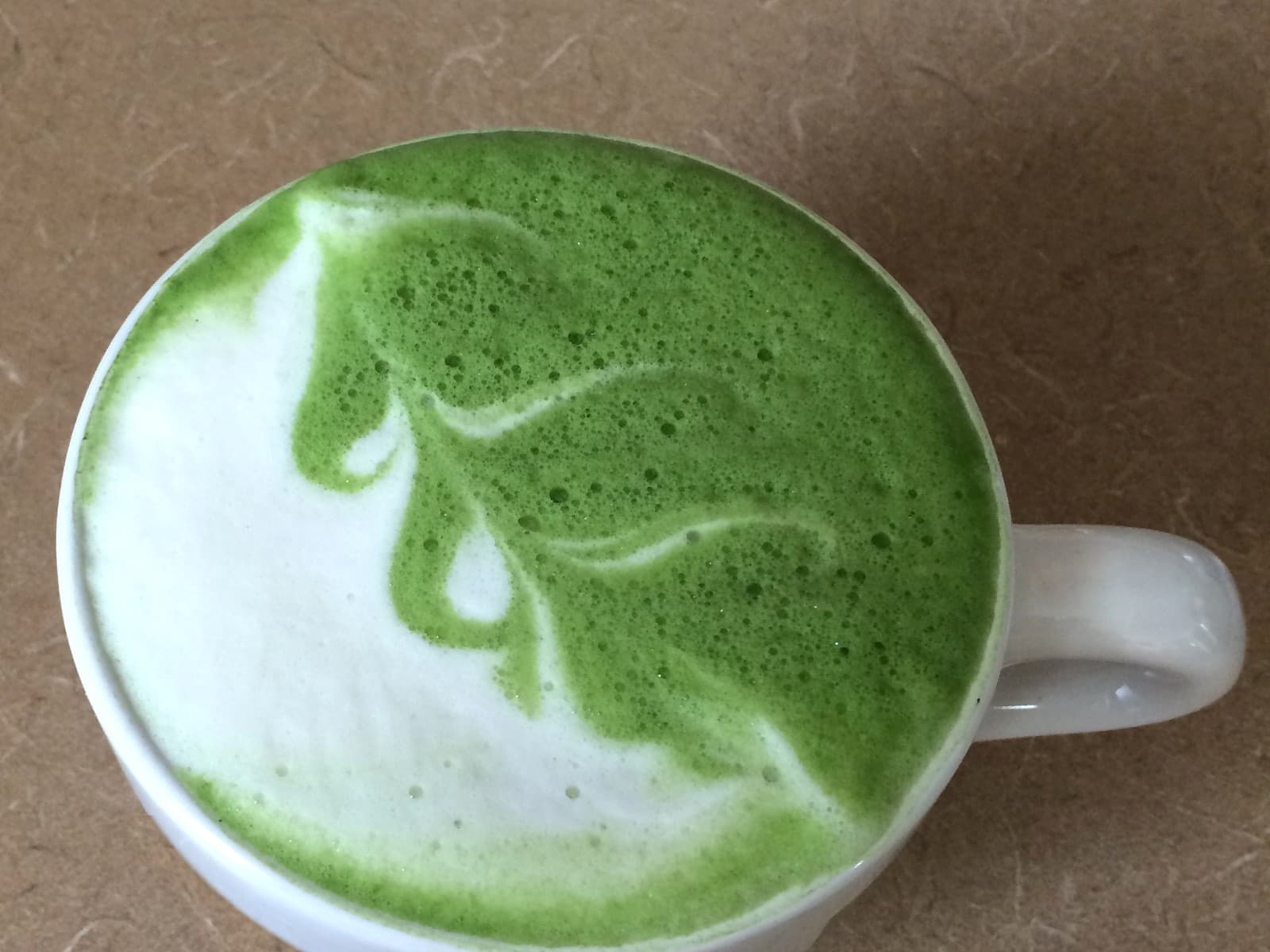 Pistachio vanilla matcha latte with a leaf-shaped swirl in the foam, in a white mug on a warm tan counter.